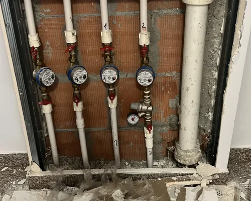 Nișă tehnică în perete cu o instalație sanitară modernă din țevi albe PPR, robineți roșii și multiple contoare de apă (apometre) montate.