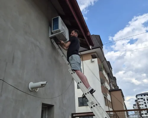 Frigotehnist lucrând pe o scară foarte înaltă pentru a fixa unitatea externă a unui aparat de aer condiționat sub streașina unei clădiri rezidențiale.