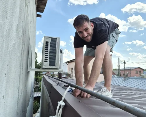 Tehnician zâmbind în timp ce lucrează pe un acoperiș metalic la trasarea și fixarea țevilor de cupru pentru un aparat de aer condiționat.