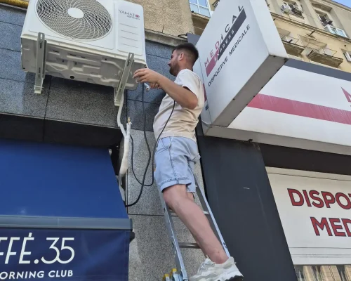 Un tehnician instalează unitatea externă a unui aparat de aer condiționat Yamato, stând pe o scară la exteriorul unei clădiri comerciale.