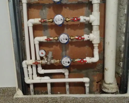 Lucrare în desfășurare la o instalație sanitară încastrată în perete, cu apometre și țevi noi, având moloz și resturi de finisaje pe podea.